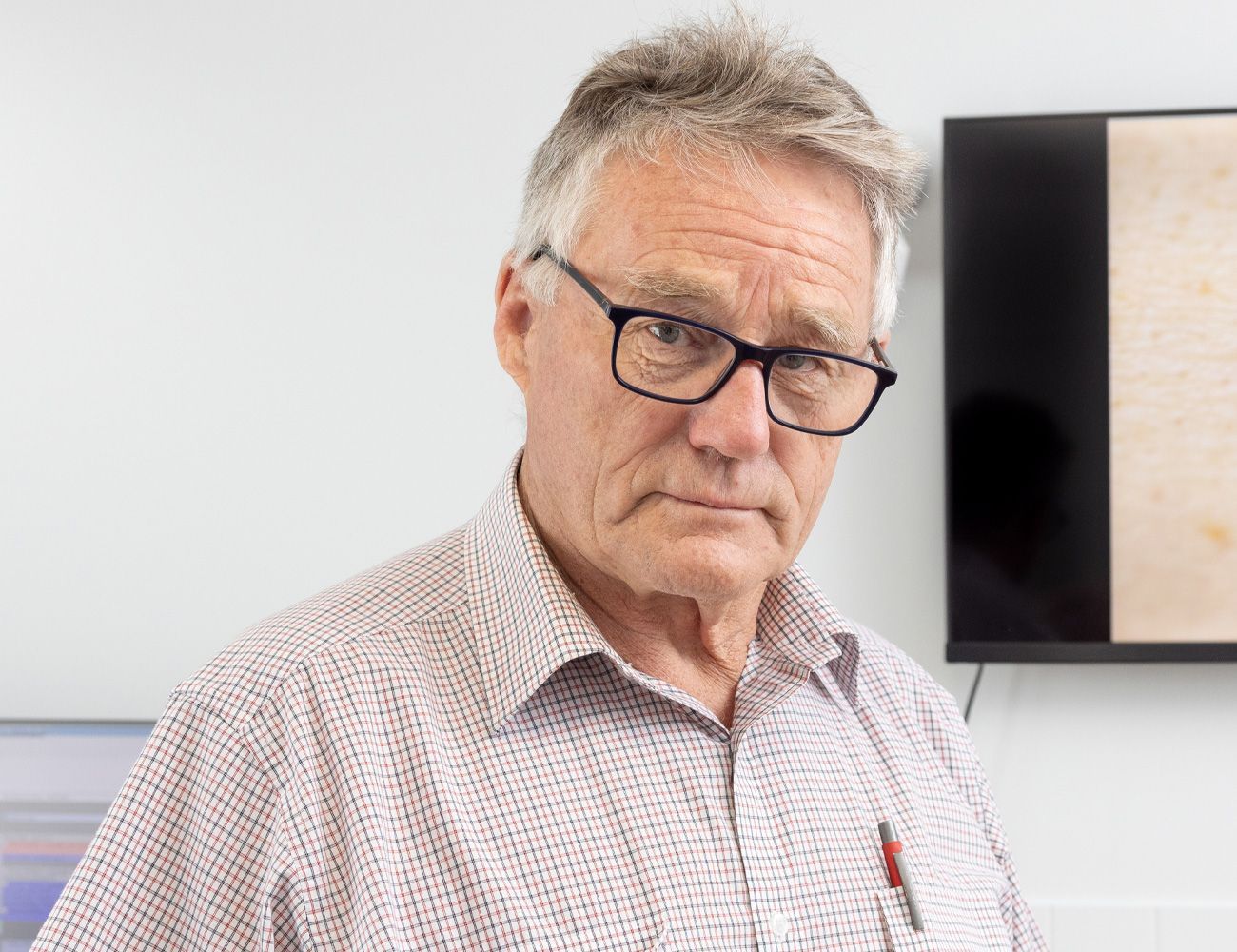 An image of Dr John Kiss wearing glasses and a pen is standing in front of a television.