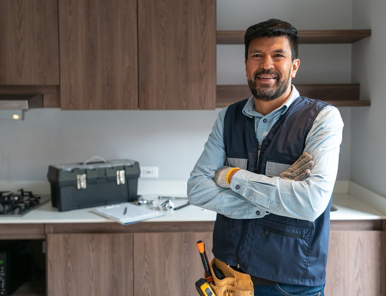 A man is standing in a kitchen with his arms crossed and holding a toolbox.