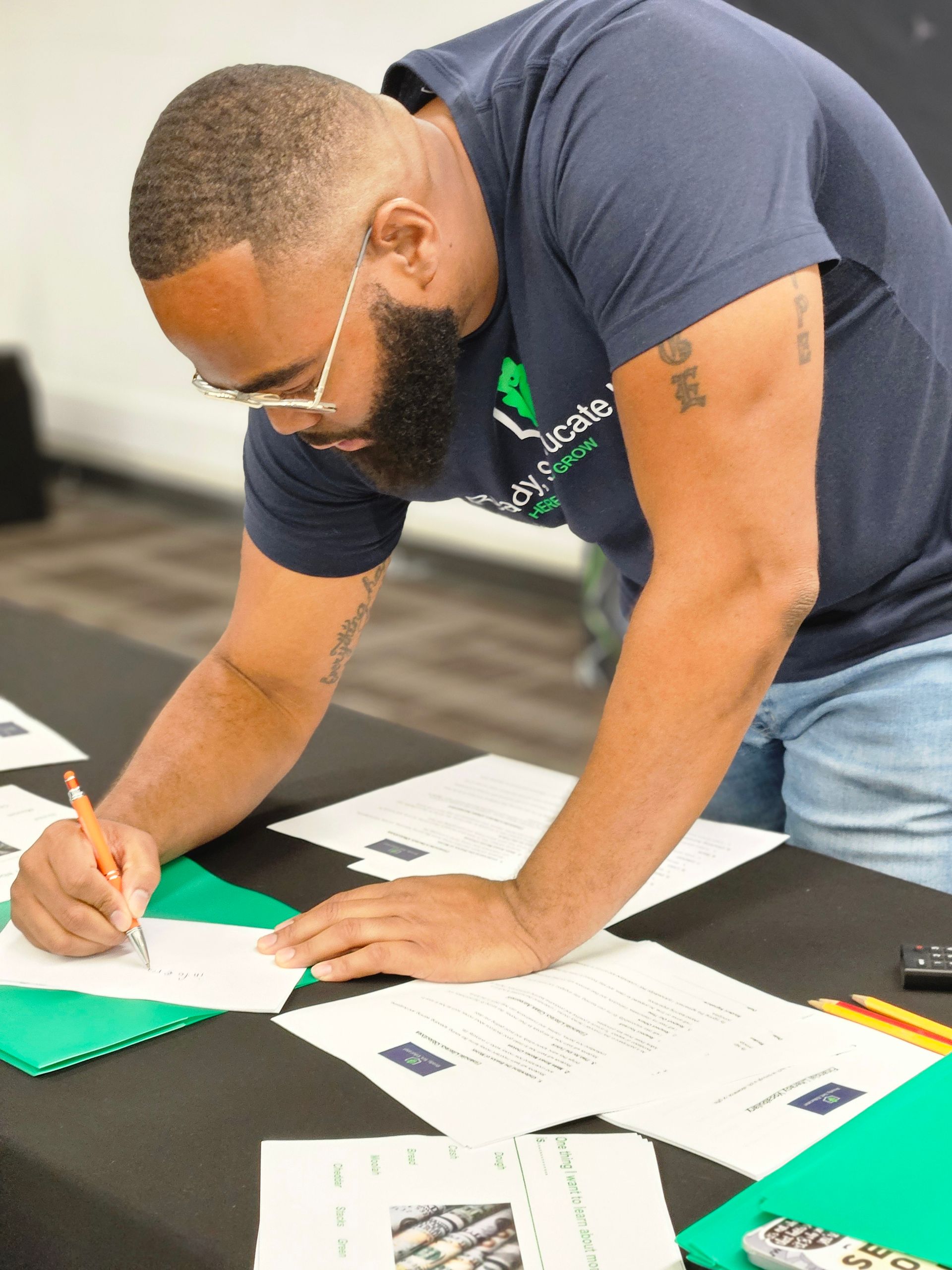 Man with beard signing documents at a table. He is wearing glasses and a blue shirt.