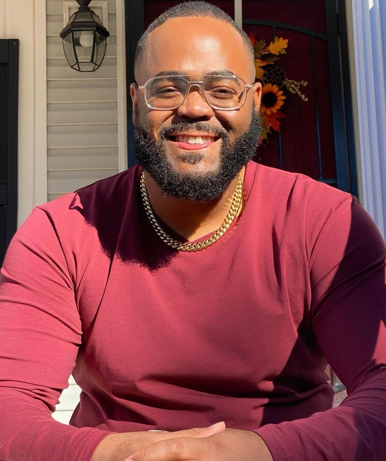 Man with a beard, wearing glasses and a maroon shirt, smiles outside.