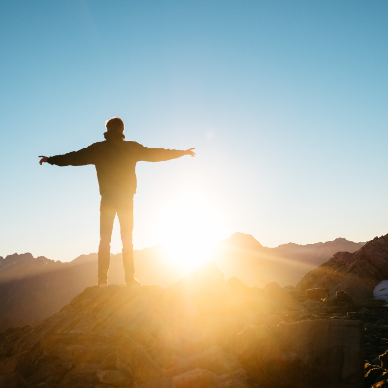 A man is standing on top of a mountain with his arms outstretched at sunset.