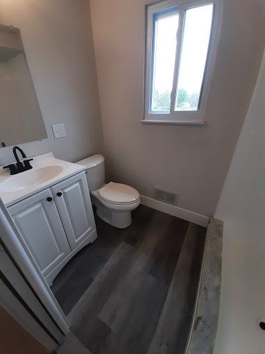 A bathroom with a toilet , sink , mirror and window.