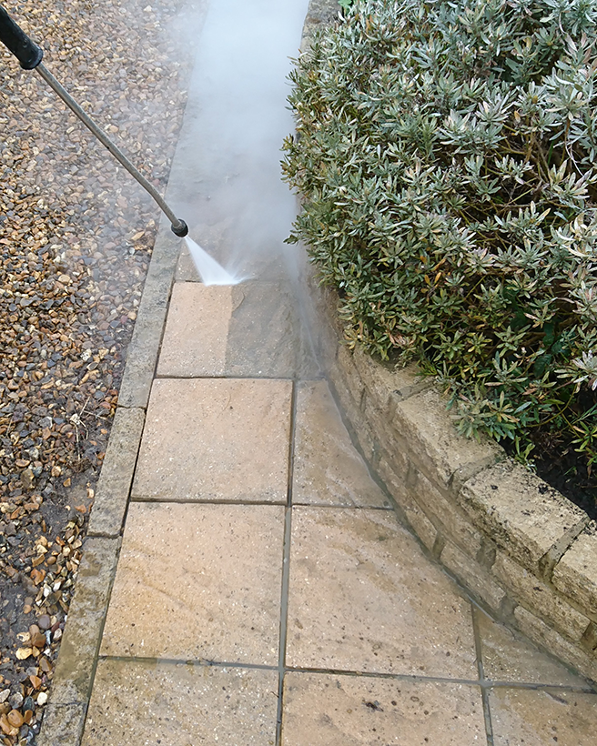 Pressure washer cleaning a stone walkway next to a green bush and brick border.