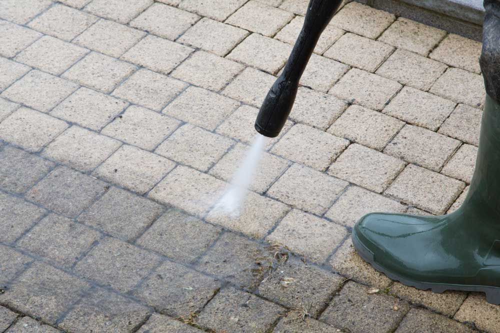 A person in green boots power washes a light-colored brick patio, cleaning the dirty surface.