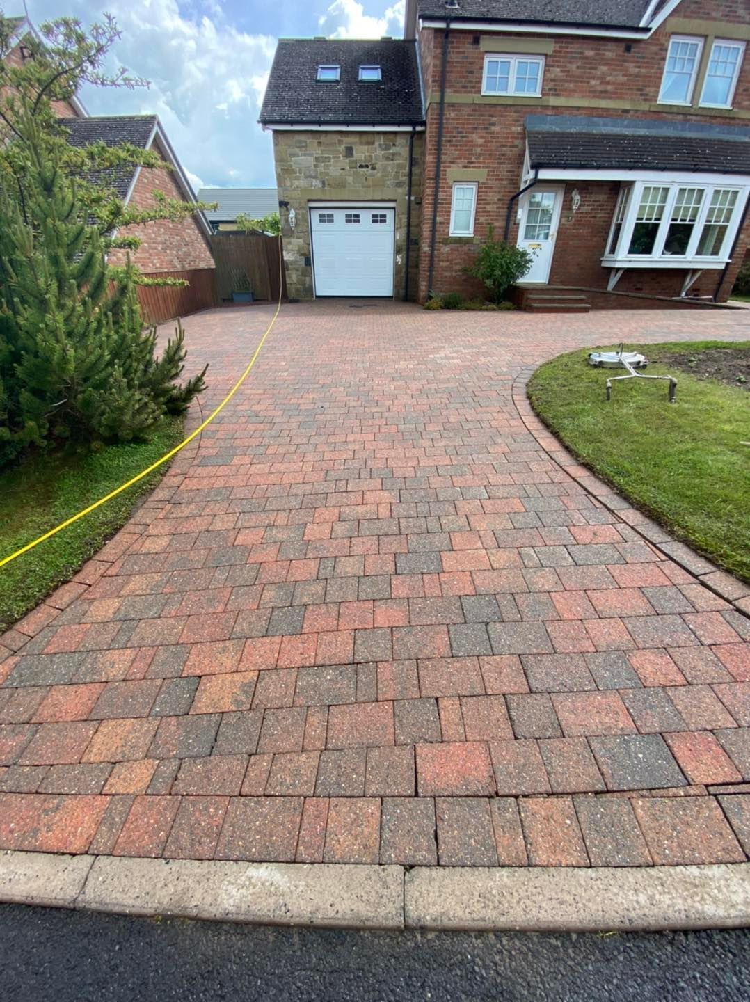 Brick driveway leading to a house with a garage. The driveway has red-brown bricks and a curved border of grass.