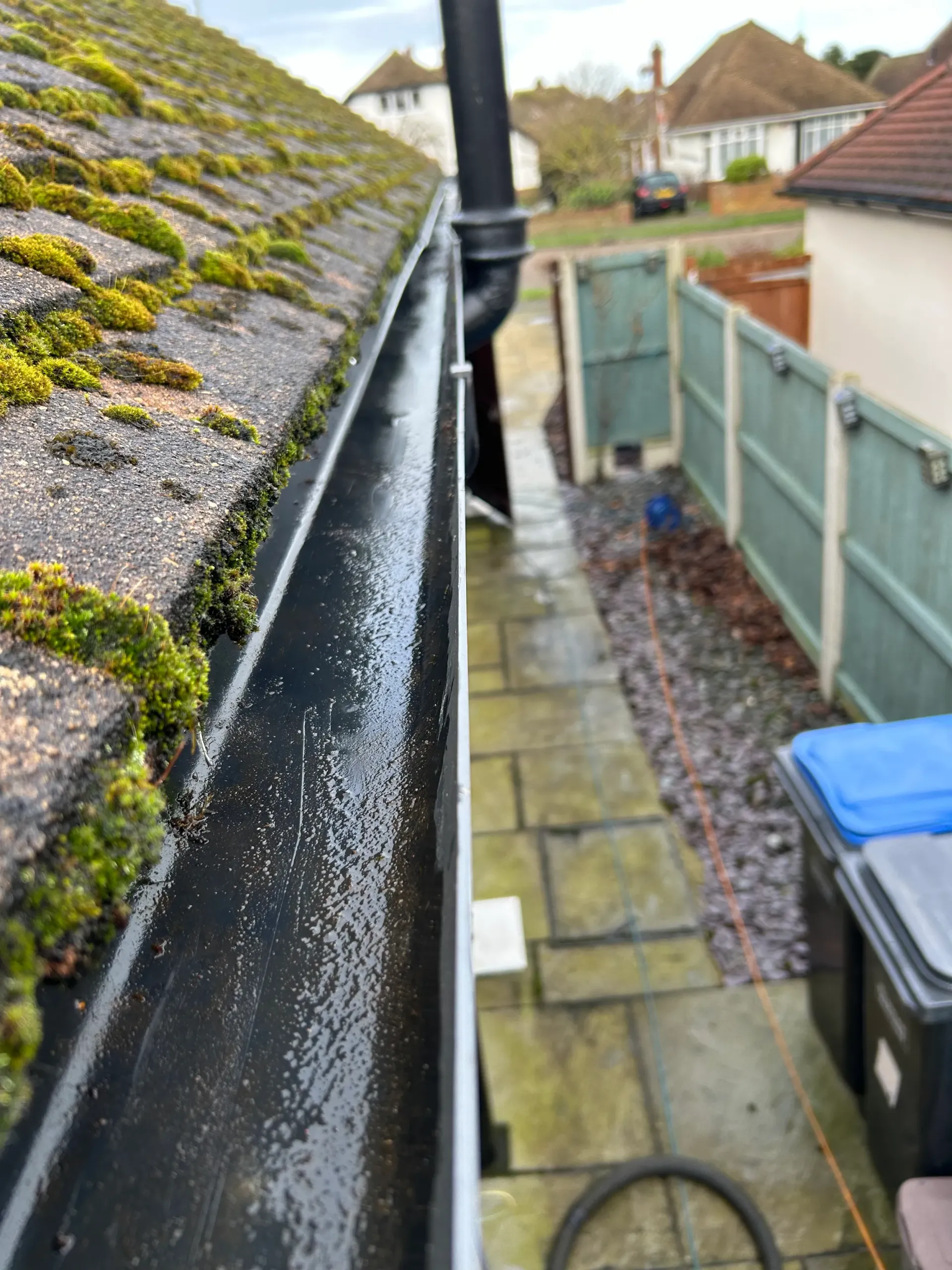 Black gutter with moss-covered roof edge, adjacent to a paved pathway and a fence.
