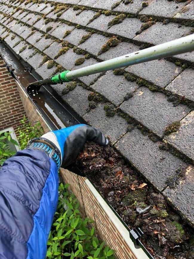 Person cleaning a gutter filled with debris; gloves visible, using a long-handled tool, outside on a roof.
