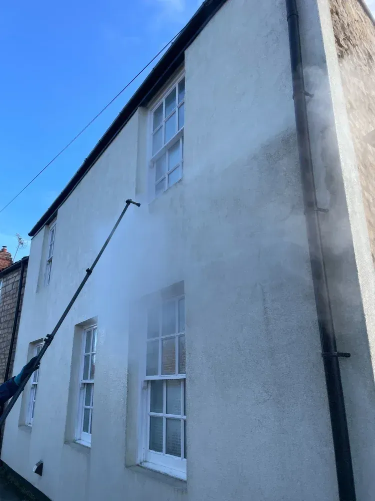 A person power washing the exterior of a white two-story building on a sunny day.