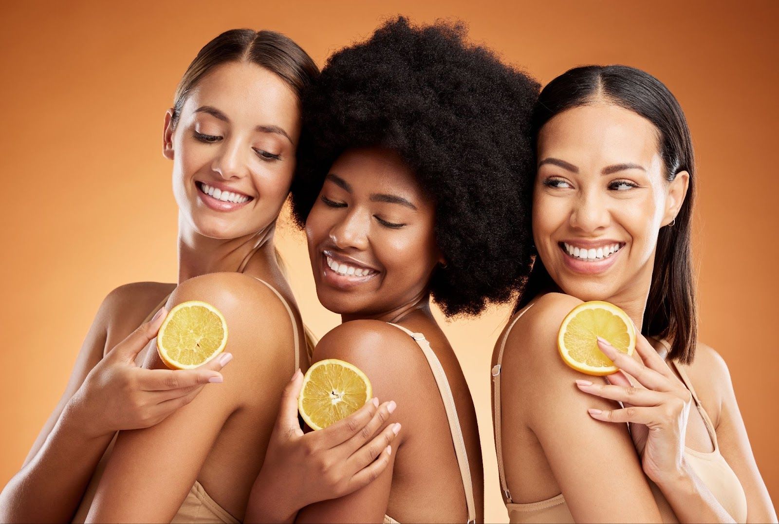 Three women smiling and holding orange slices against their shoulders on an orange background.