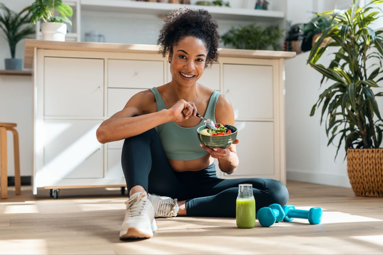 Woman in workout clothes eating a healthy bowl with a smoothie and dumbbells nearby.