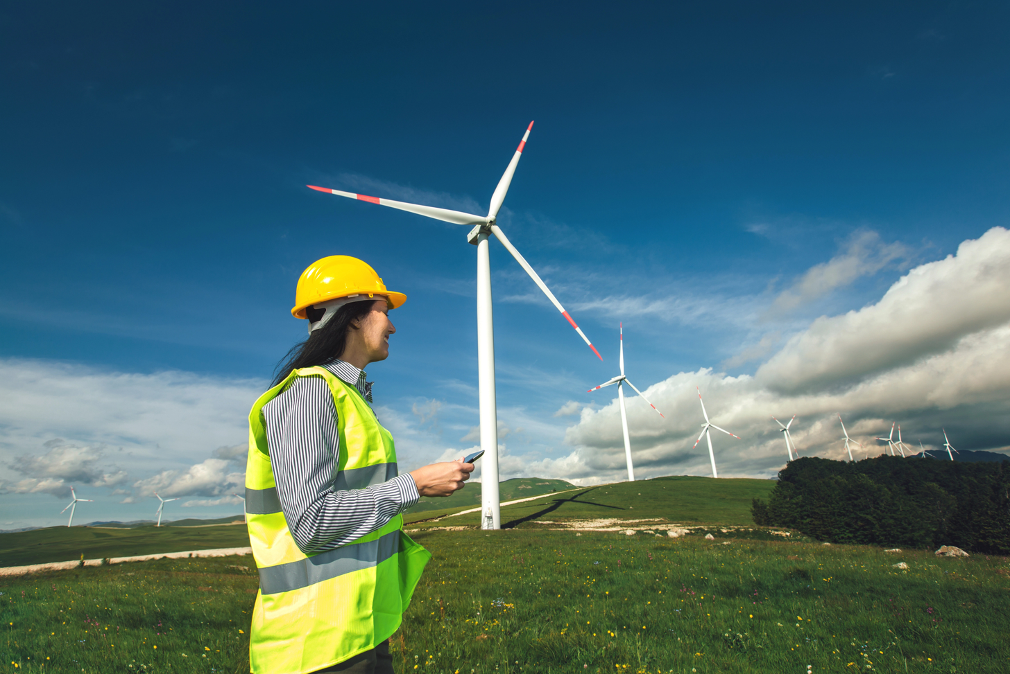 Una mujer con equipo de seguridad inspecciona las turbinas eólicas en un campo bajo un cielo azul.