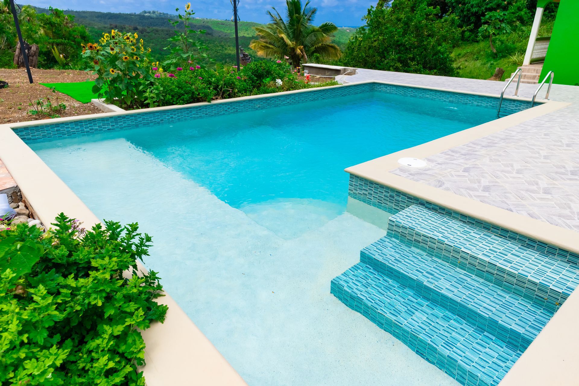 Blue tiled swimming pool with steps, surrounded by a concrete patio and lush green plants, with a hillside backdrop.