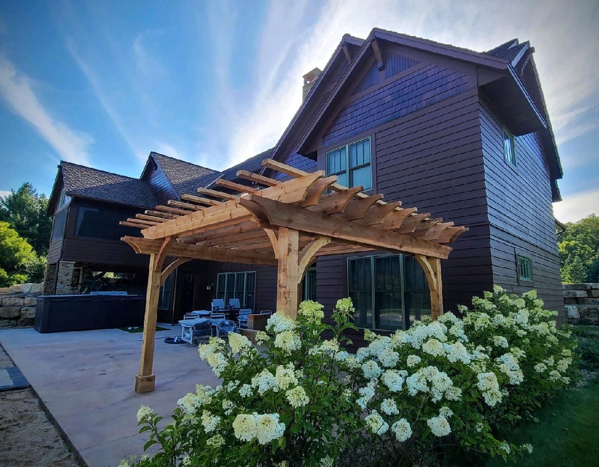 Wooden pergola attached to a brown house with a blooming white rose bush. Sunny day.