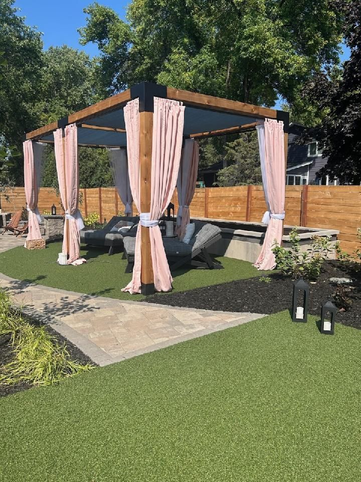 Pergola with pink drapes over outdoor seating area on artificial turf, with a paved walkway and wooden fence.