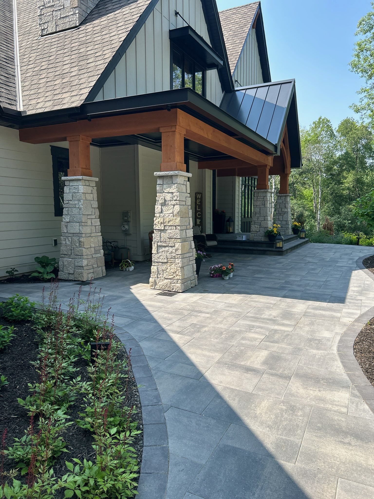 Stone and wood porch with paved walkway leading to a house entrance. Green foliage surrounds the walkway.