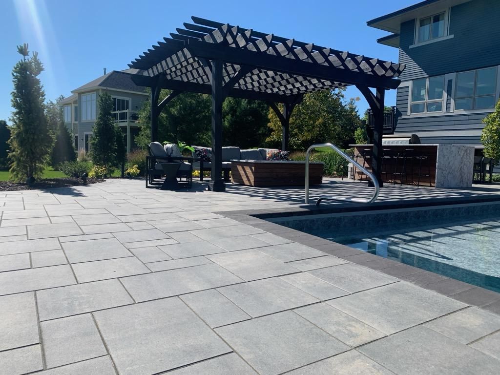 Patio with pergola, pool, and seating area. Grey pavers, blue sky, and a sunny day.