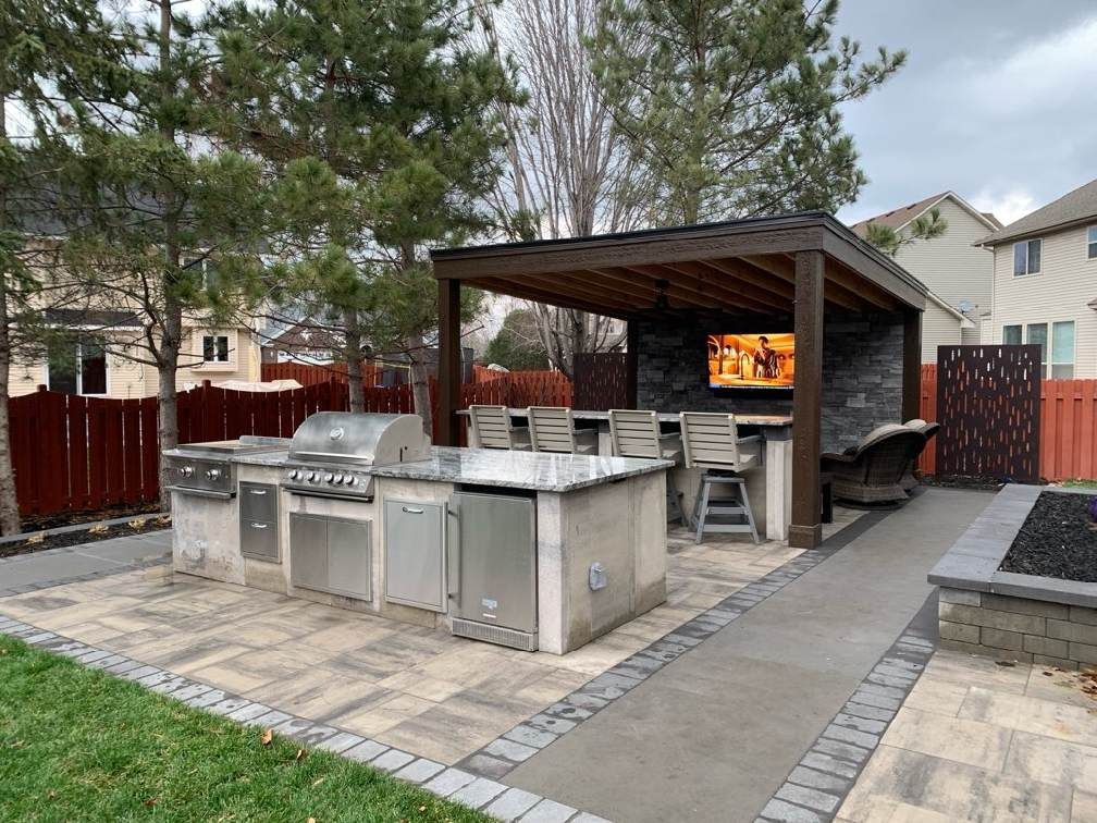 Outdoor kitchen with grill, countertop, seating, and a TV under a pergola.