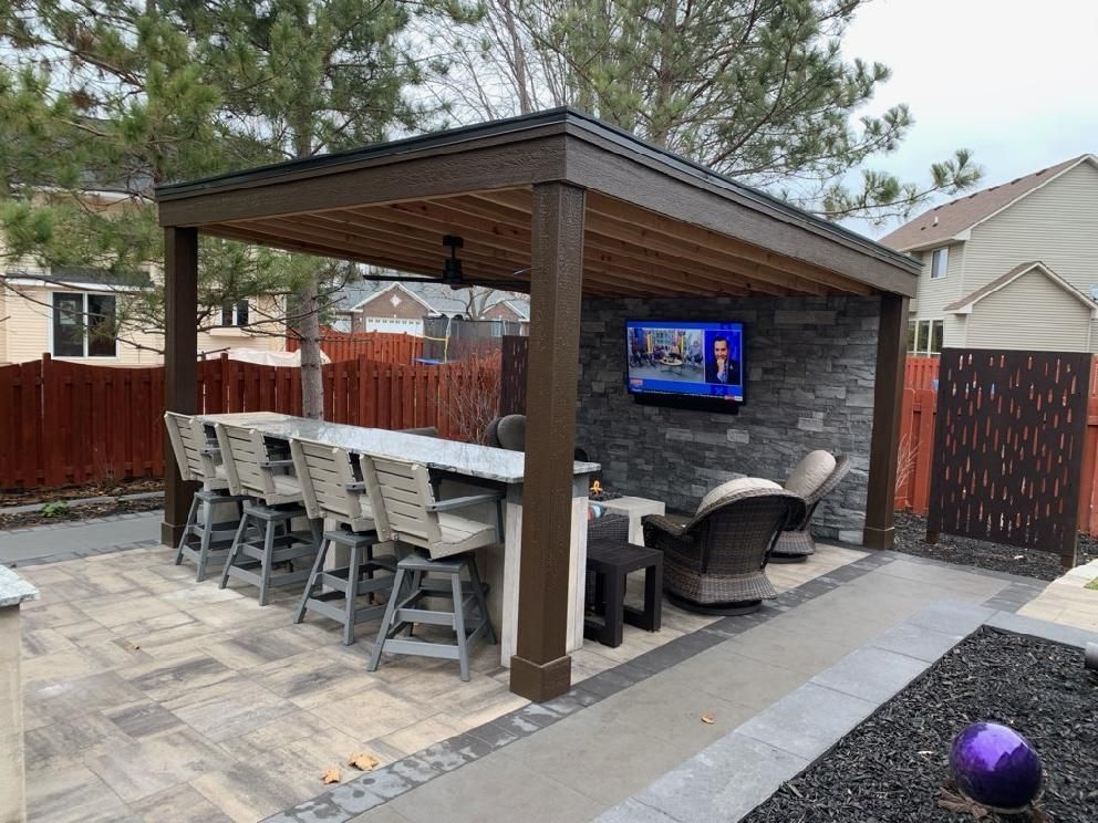 Outdoor bar with stone wall, TV, seating, and long countertop with bar stools under a pergola roof.
