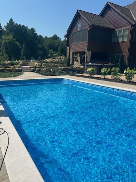 Swimming pool with blue water next to a two-story brown house, stone patio, and landscaped yard.
