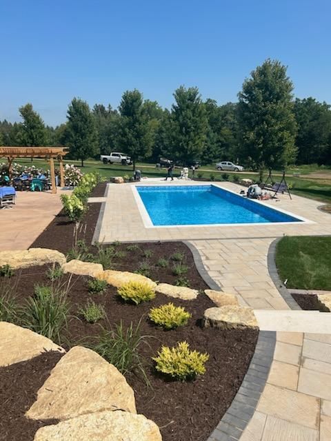 Rectangular pool with blue water, surrounded by patio and landscaped with rocks and greenery, under a blue sky.