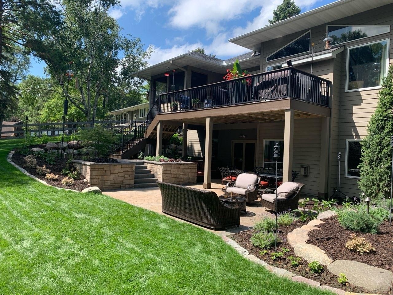 Backyard with a two-story deck, patio, and lush green lawn. Outdoor seating, landscaping, and a sunny sky are visible.