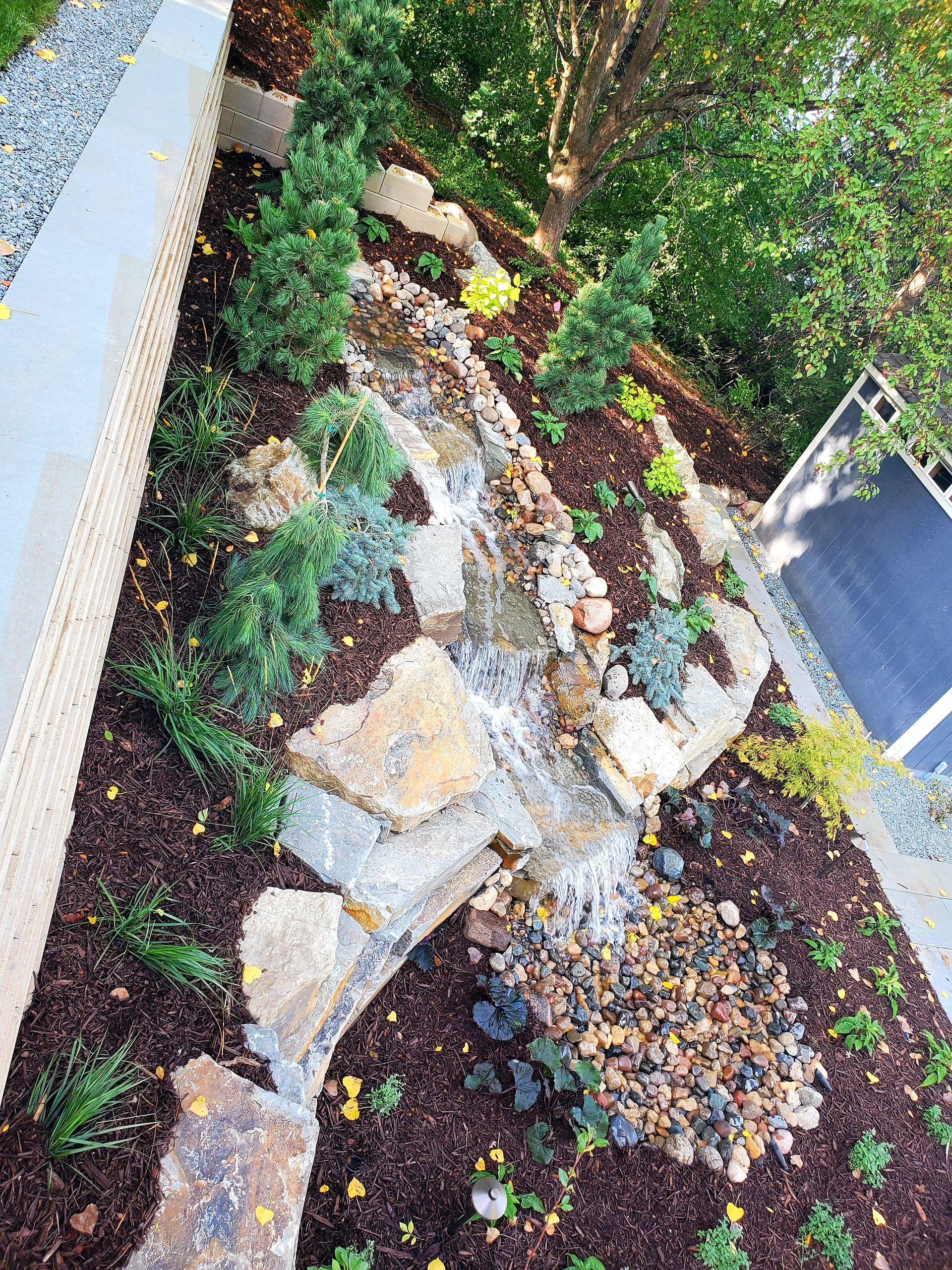 A landscaped garden with a small waterfall, rocks, and various plants. Brown mulch and green foliage.