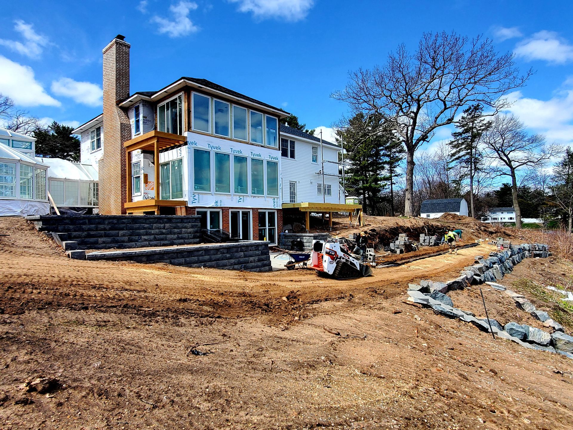 House under construction with dirt ground, retaining wall, and blue sky.