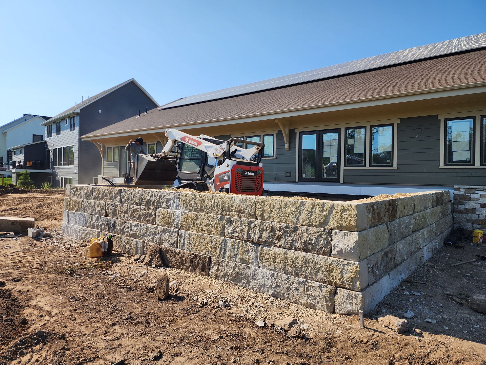 A small excavator on a retaining wall made of beige blocks, next to a house with a green facade, under a clear sky.
