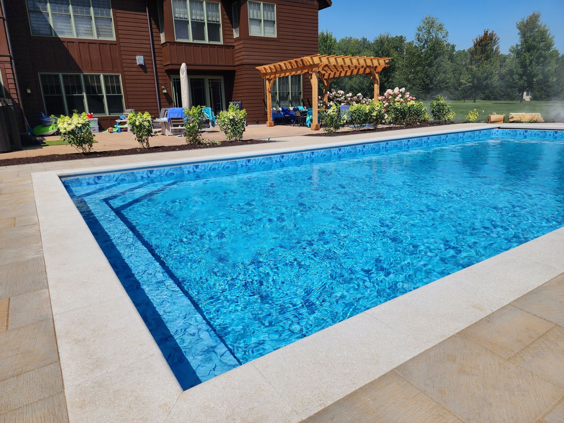 A blue, rectangular swimming pool with steps, bordered by light-colored stone, in front of a brown house.