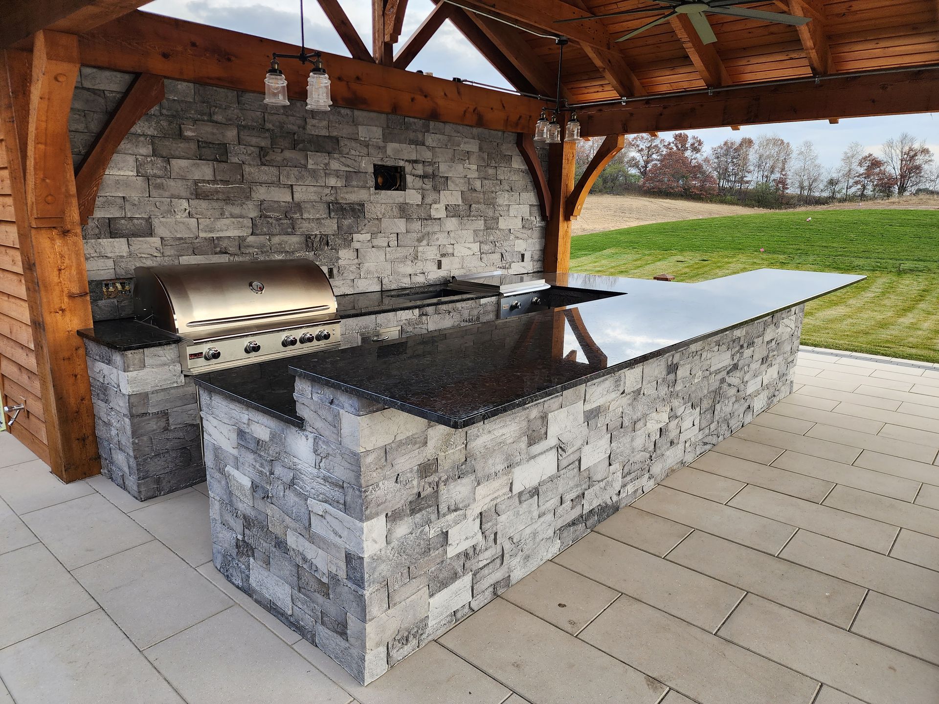 Outdoor kitchen with stone counters, grill, and wood-beam roof, with a grassy field in the background.