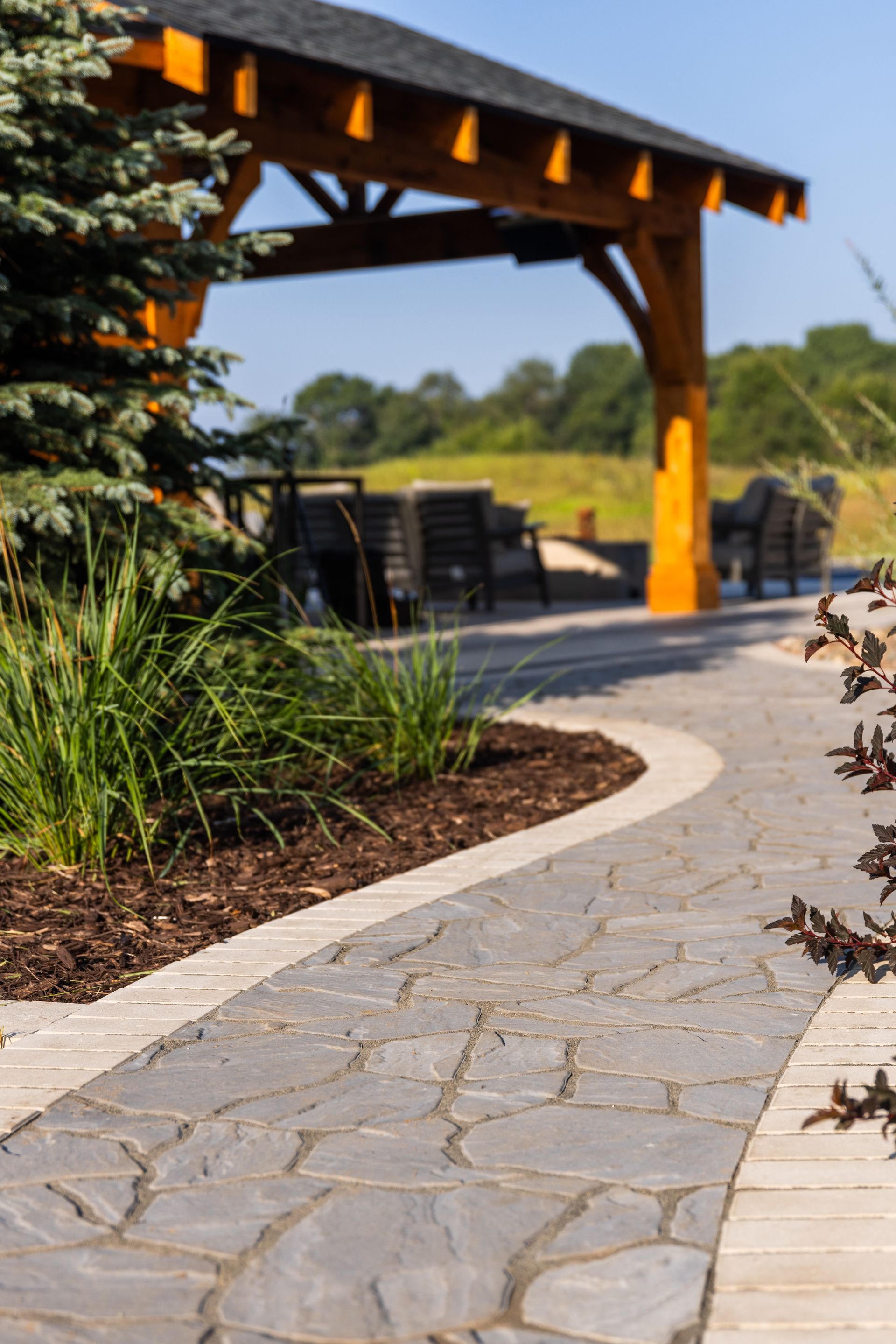 Stone path curves toward a wooden gazebo with outdoor seating. Lush greenery borders path.