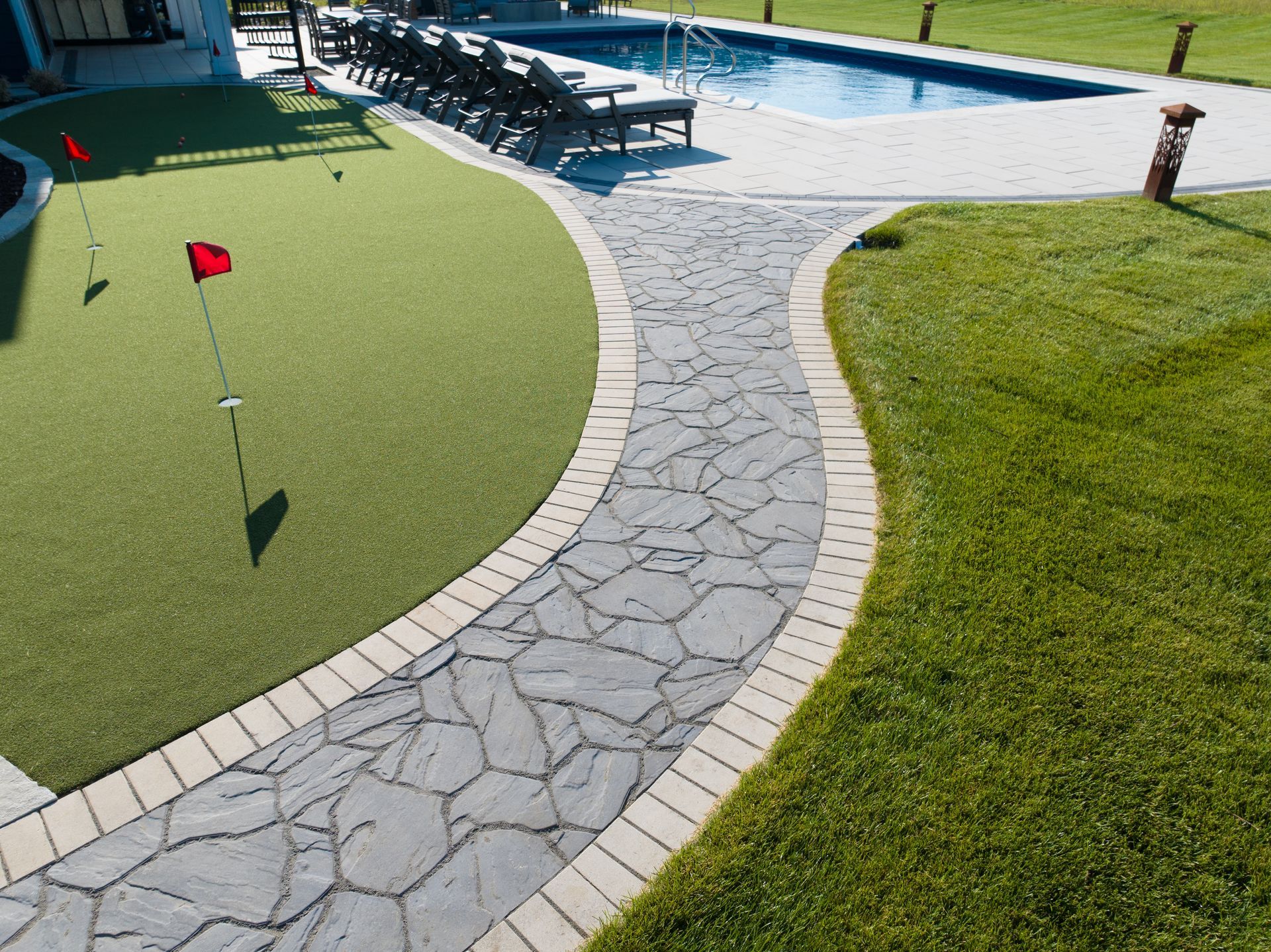 Putting green and stone pathway curving around a pool, bordered by grass and tan bricks.