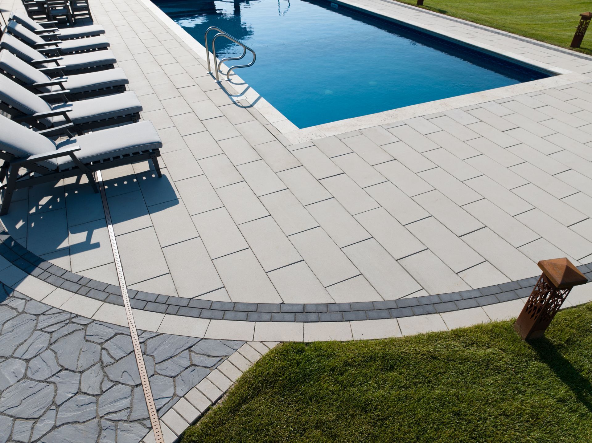 Poolside scene: rectangular pool with blue water, gray pavers, lounge chairs, and green grass.