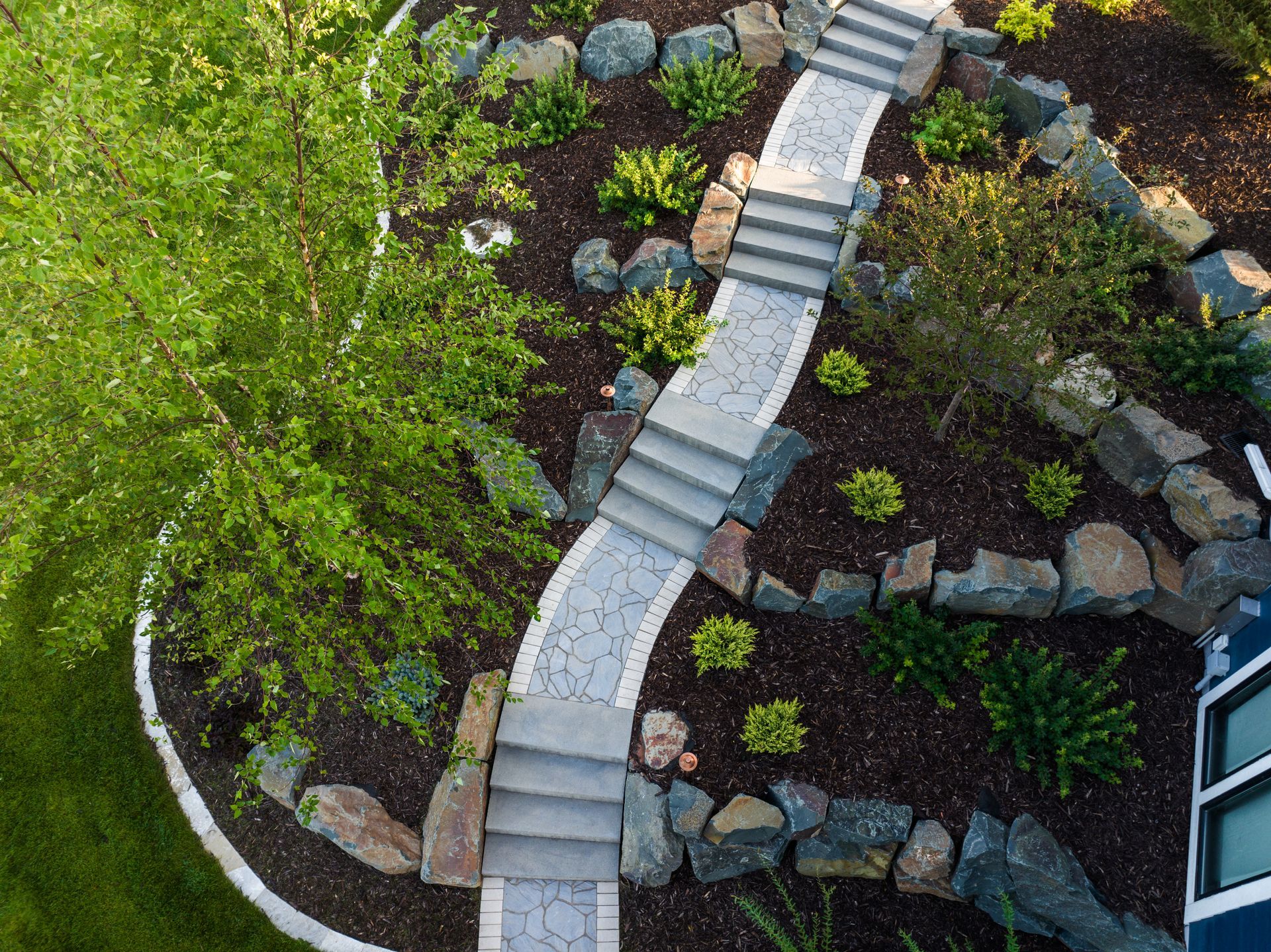 Aerial view of a garden with a winding stone pathway, steps, and rock borders, surrounded by plants and trees.