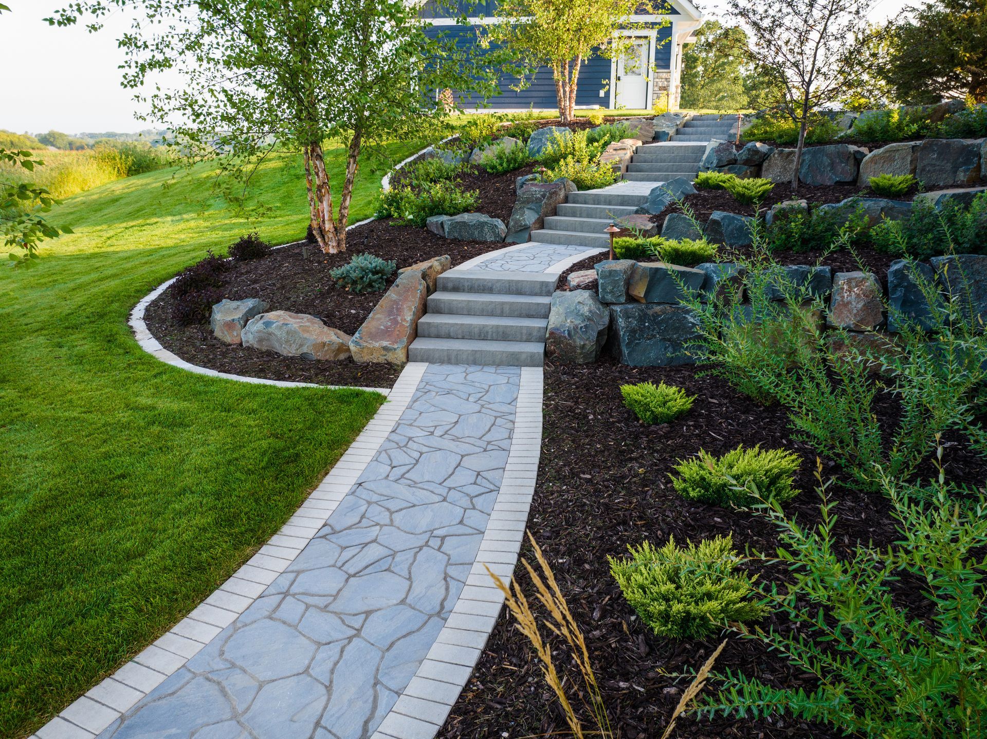 Stone path and steps lead up a hillside to a blue building; landscaped with rocks, plants, and green grass.
