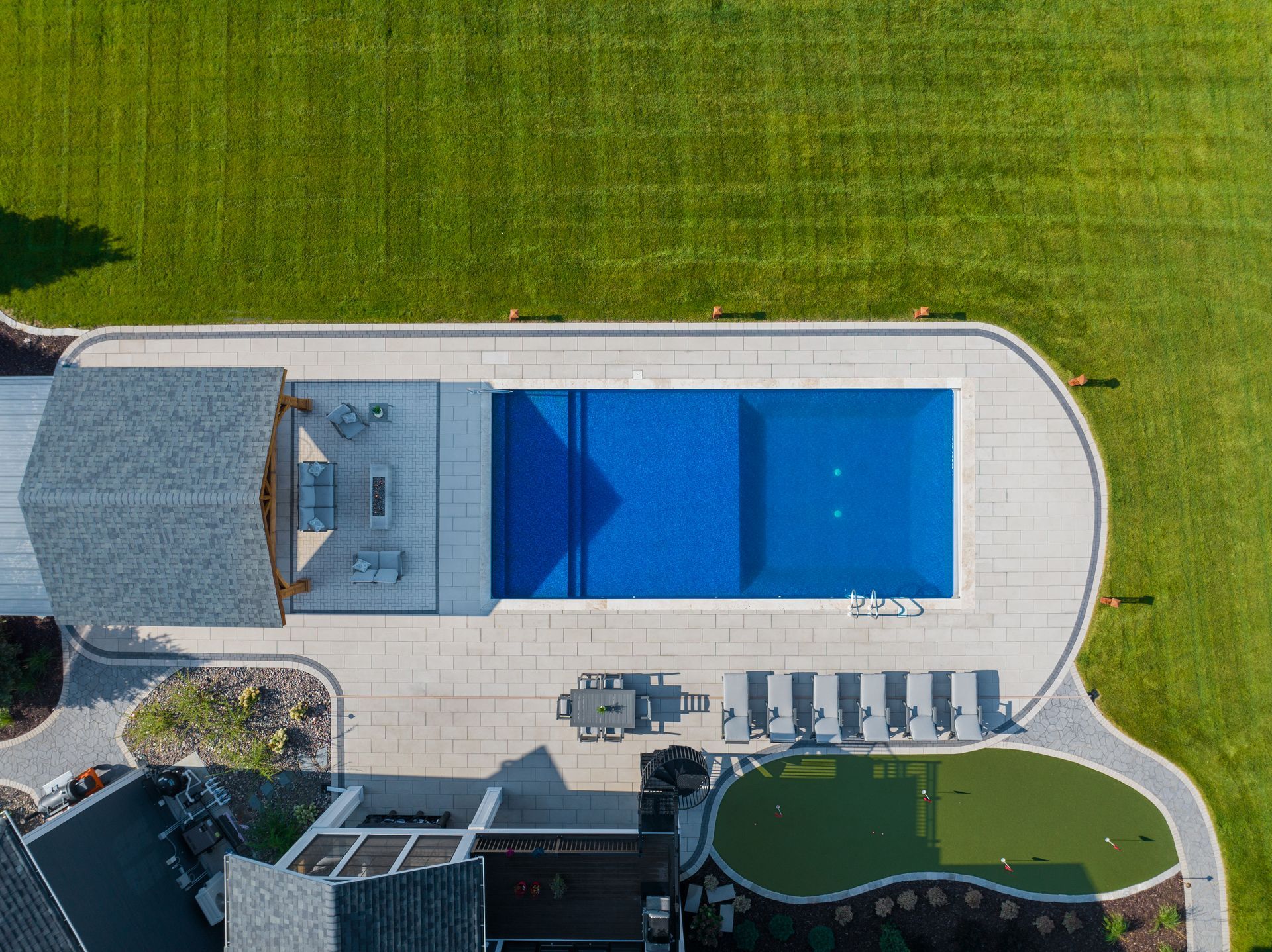 Overhead view of a rectangular blue pool surrounded by a patio, lawn, and a small putting green.