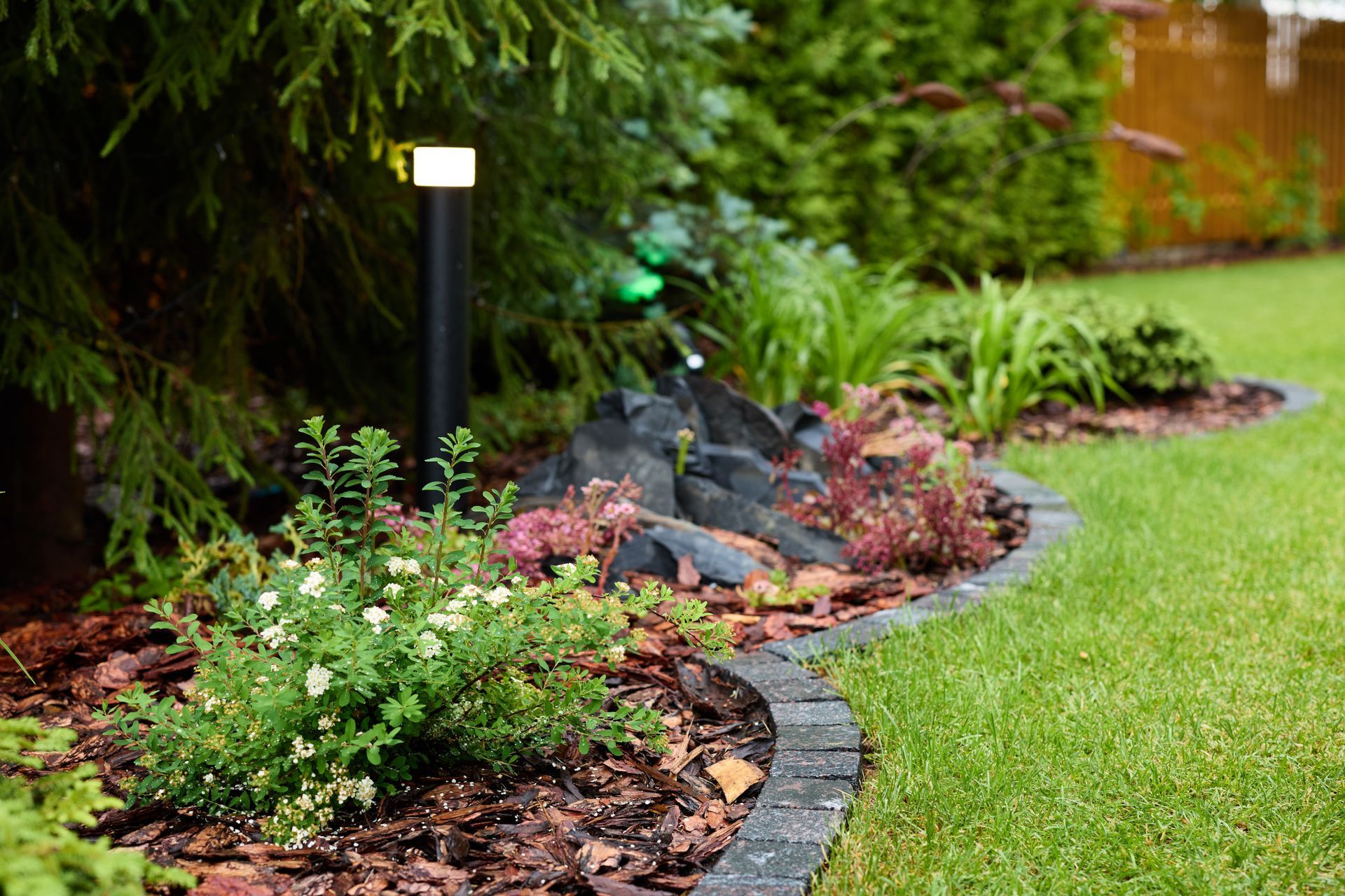 Well-lit garden bed with dark edging, rocks, greenery, and a tall black lamp next to grass.