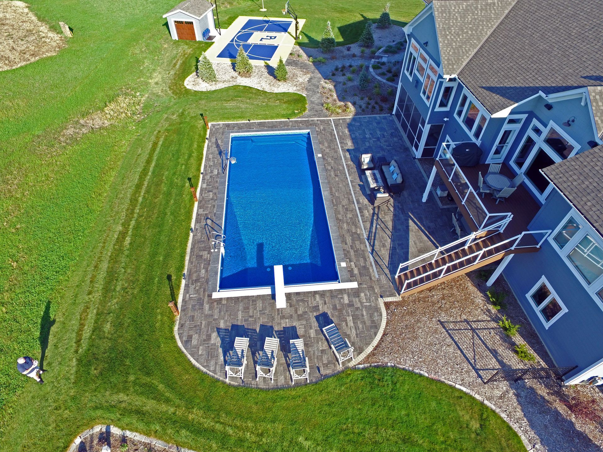 Aerial view of a rectangular pool with diving board, surrounded by patio and lawn, adjacent to a blue house.