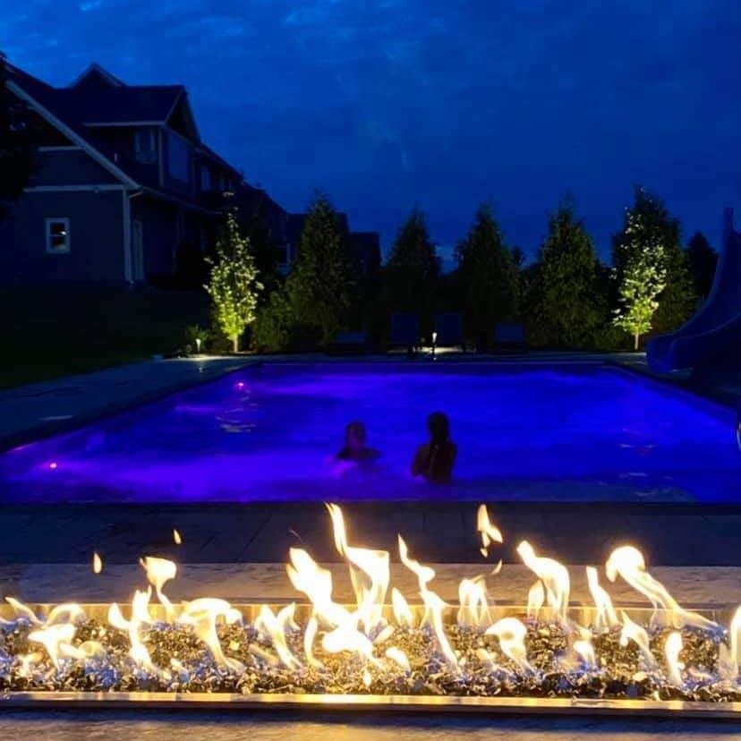 Pool at night with two people swimming, illuminated by blue lights, and a fire pit in foreground.