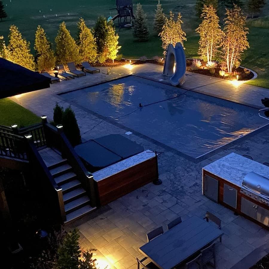 Nighttime backyard scene with a pool, hot tub, and outdoor kitchen illuminated by warm lighting.
