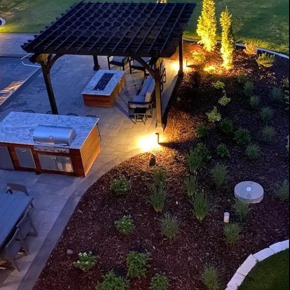 Night view of a backyard patio with pergola, outdoor kitchen, fire pit, and landscaping with illuminated features.