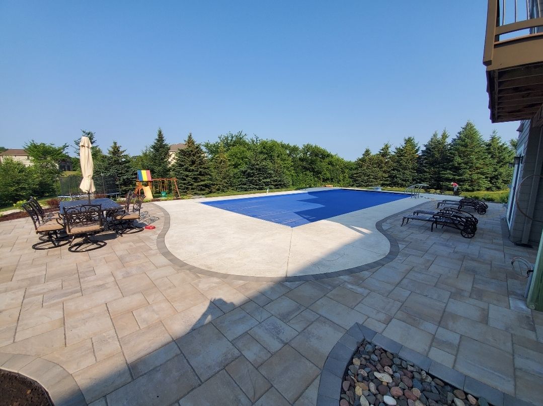Backyard patio with a blue-covered pool, dining set, and greenery under a clear blue sky.