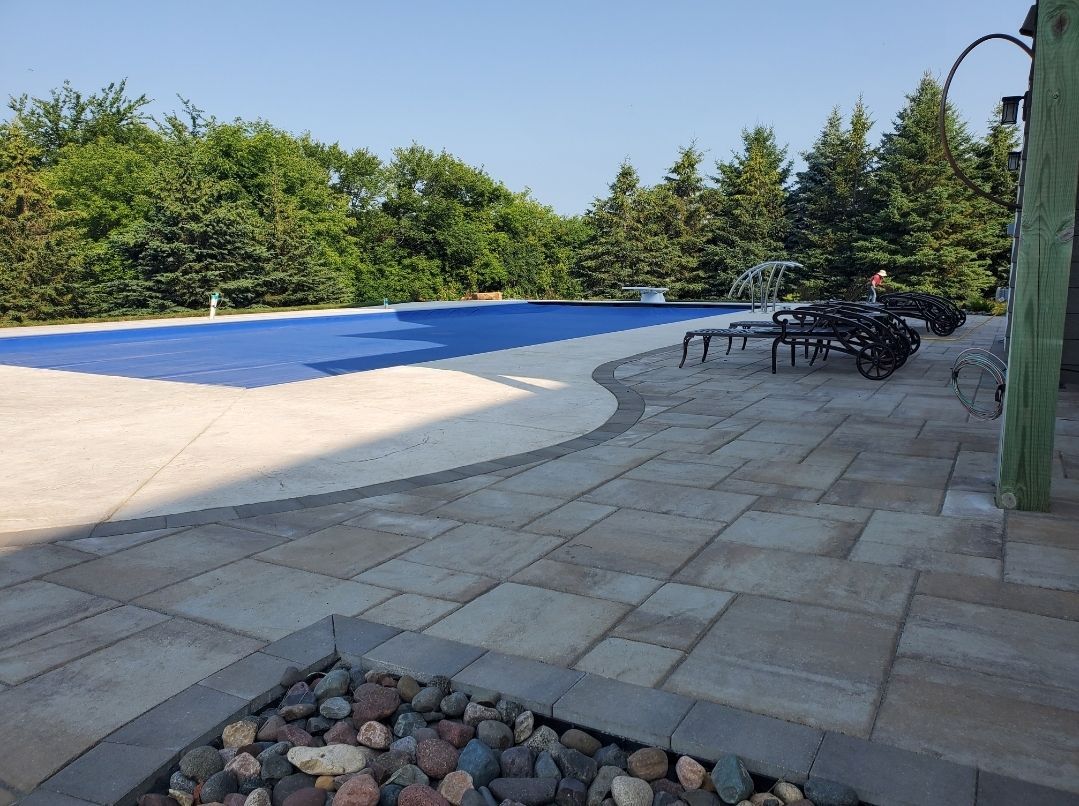 Patio with paved stones, pool with blue cover, trees, and lounge chairs. Blue sky overhead.