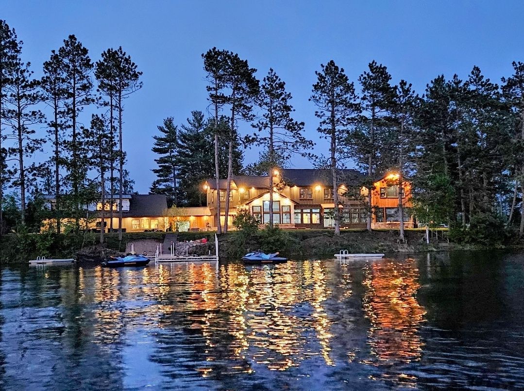 Large illuminated lakeside house at dusk, reflections in water.