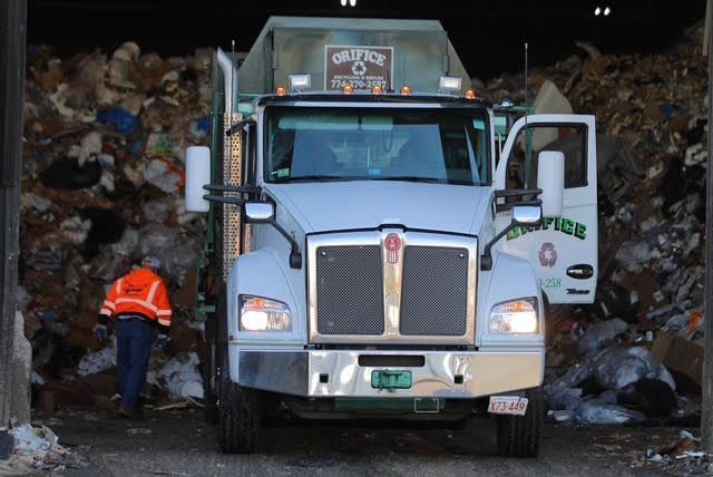 Orifice Garbage truck unloading trash at a facility; worker in orange vest.