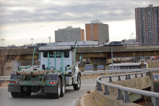 Orifice White truck on a curved road, approaching a bridge. Buildings and cloudy sky in the background.