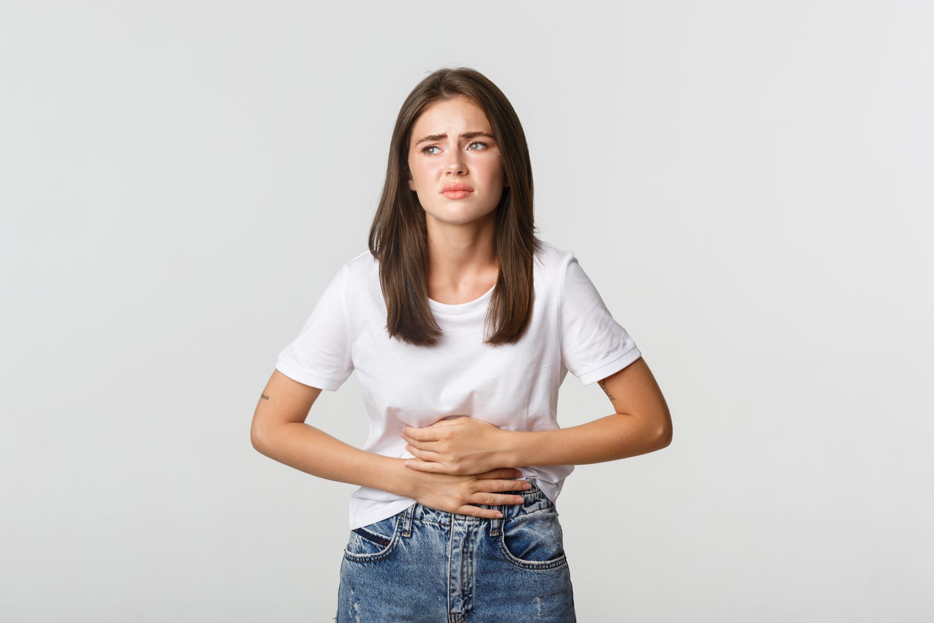 Woman in white shirt and jeans holding her stomach, looking distressed.