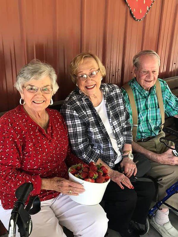 Three people smiling, holding bowl of strawberries. Outdoors, red wall.