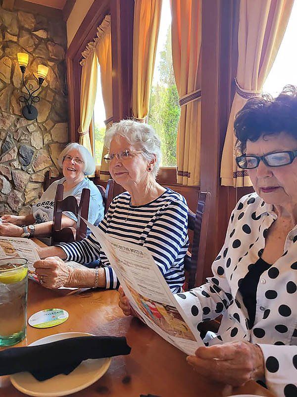 Three women at a restaurant table, looking at menus. One has a white polka dot jacket, another a striped shirt. Sunny interior.