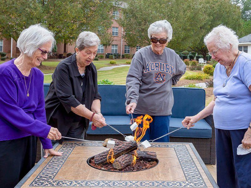 Four older women roast marshmallows over a fire pit outside, smiling, enjoying themselves.